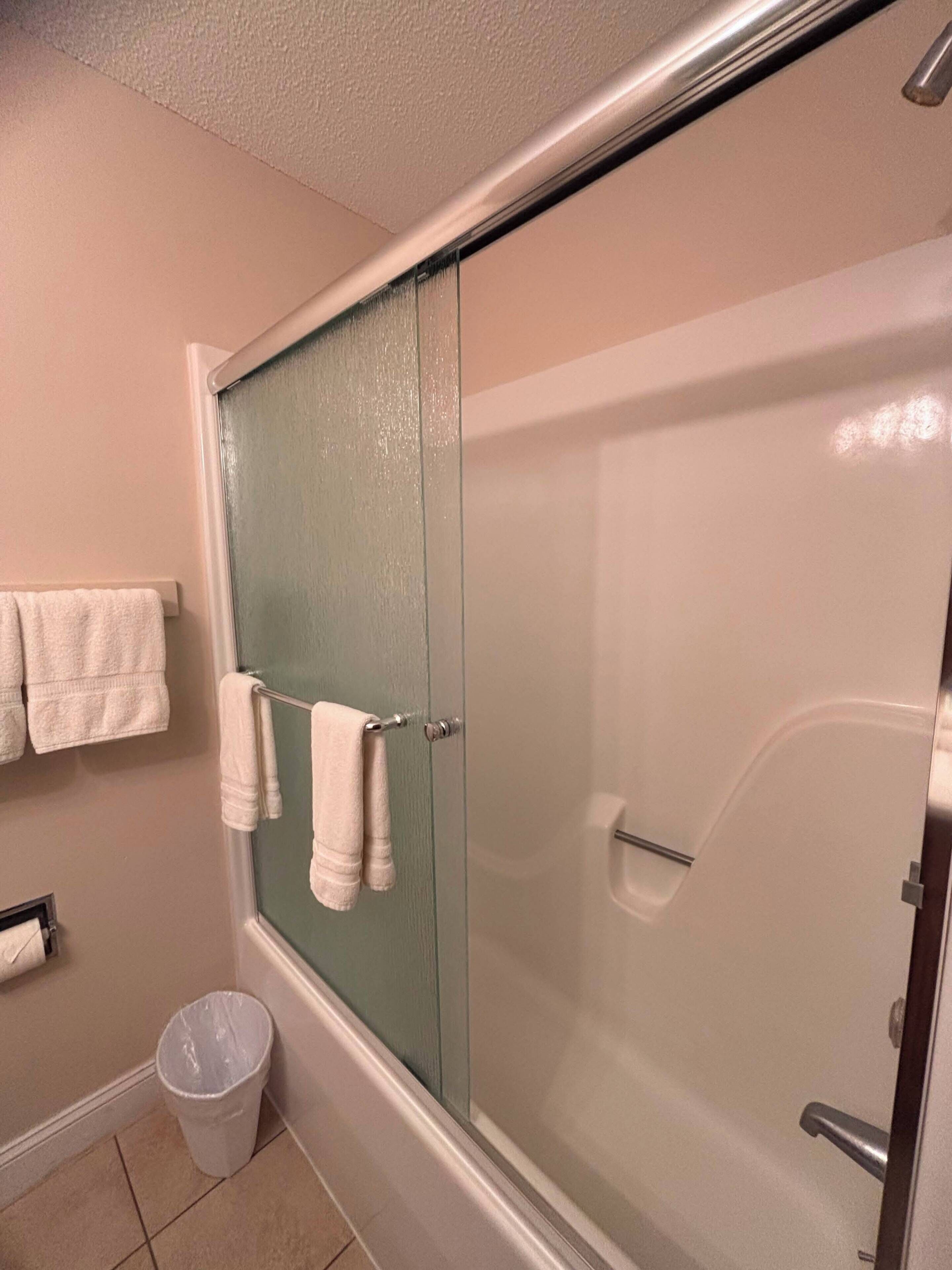 A bathroom with a glass shower door, a towel rack with two towels, a trash can, and a toilet paper holder.