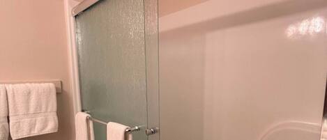 A bathroom with a glass shower door, a towel rack with two towels, a trash can, and a toilet paper holder.
