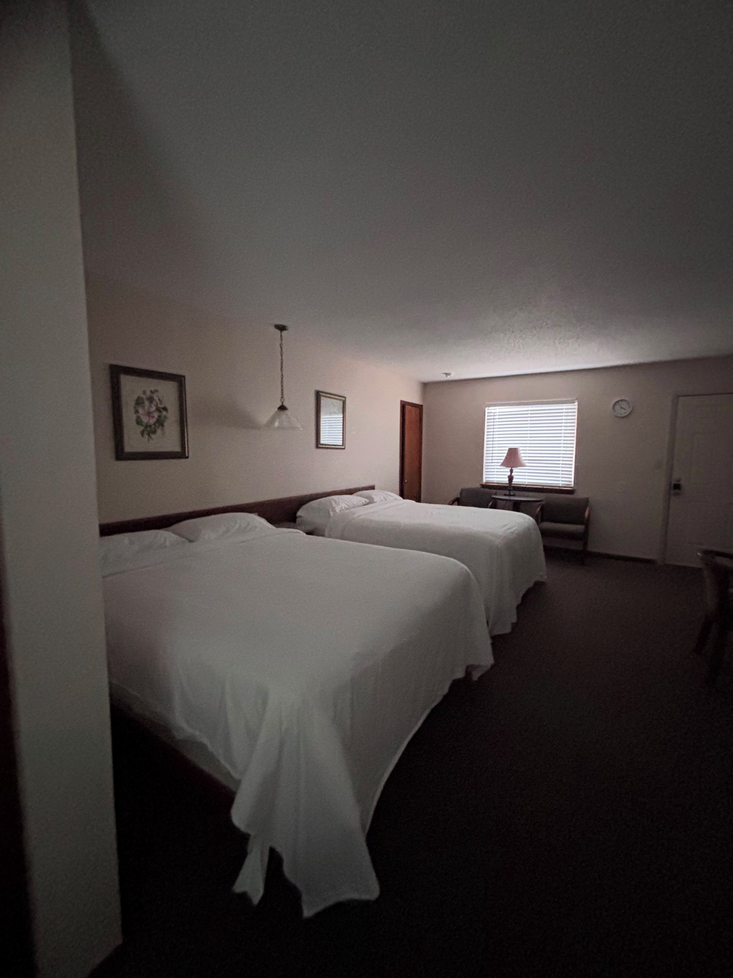 Habitación de hotel con dos camas, una mesita, una silla, una lámpara y una ventana con persianas.