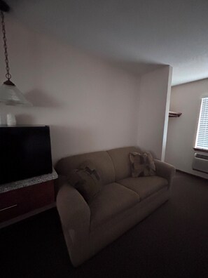 Una sala de estar con un sofá color beige, un televisor de pantalla plana sobre un mueble de madera, una lámpara colgante y una ventana con persianas.