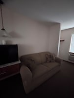 Uma sala de estar com um sofá bege, uma televisão de tela plana sobre um móvel de madeira, um lustre pendurado e uma janela com persianas.