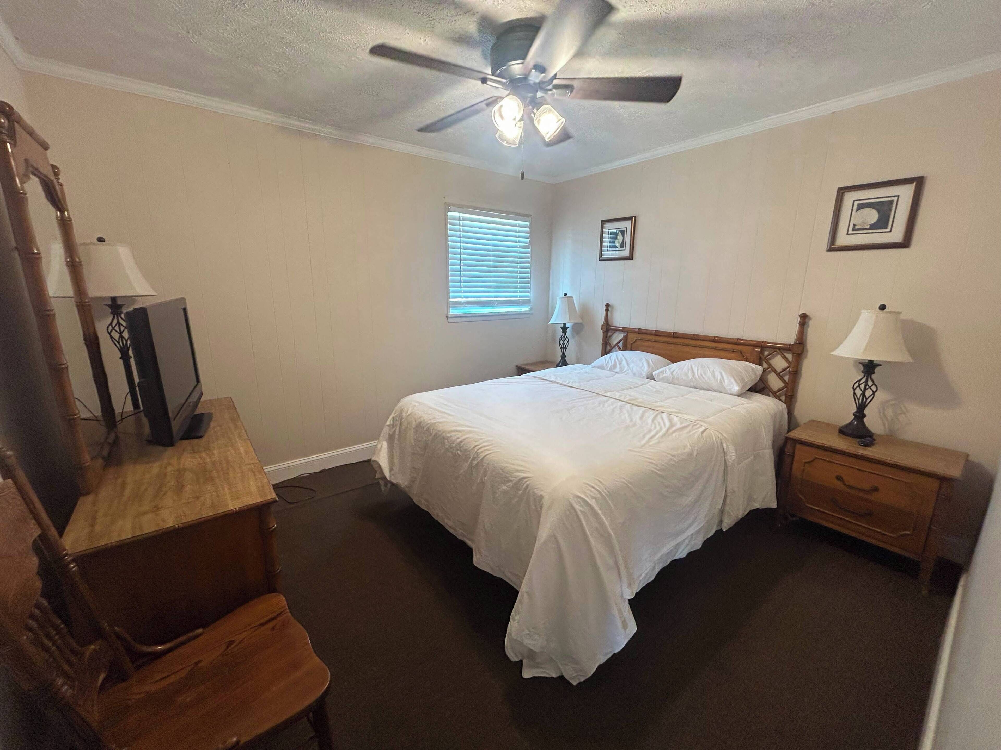 Une chambre à coucher comprenant un lit, un bureau, une chaise, un ventilateur de plafond et une table de chevet avec une lampe.