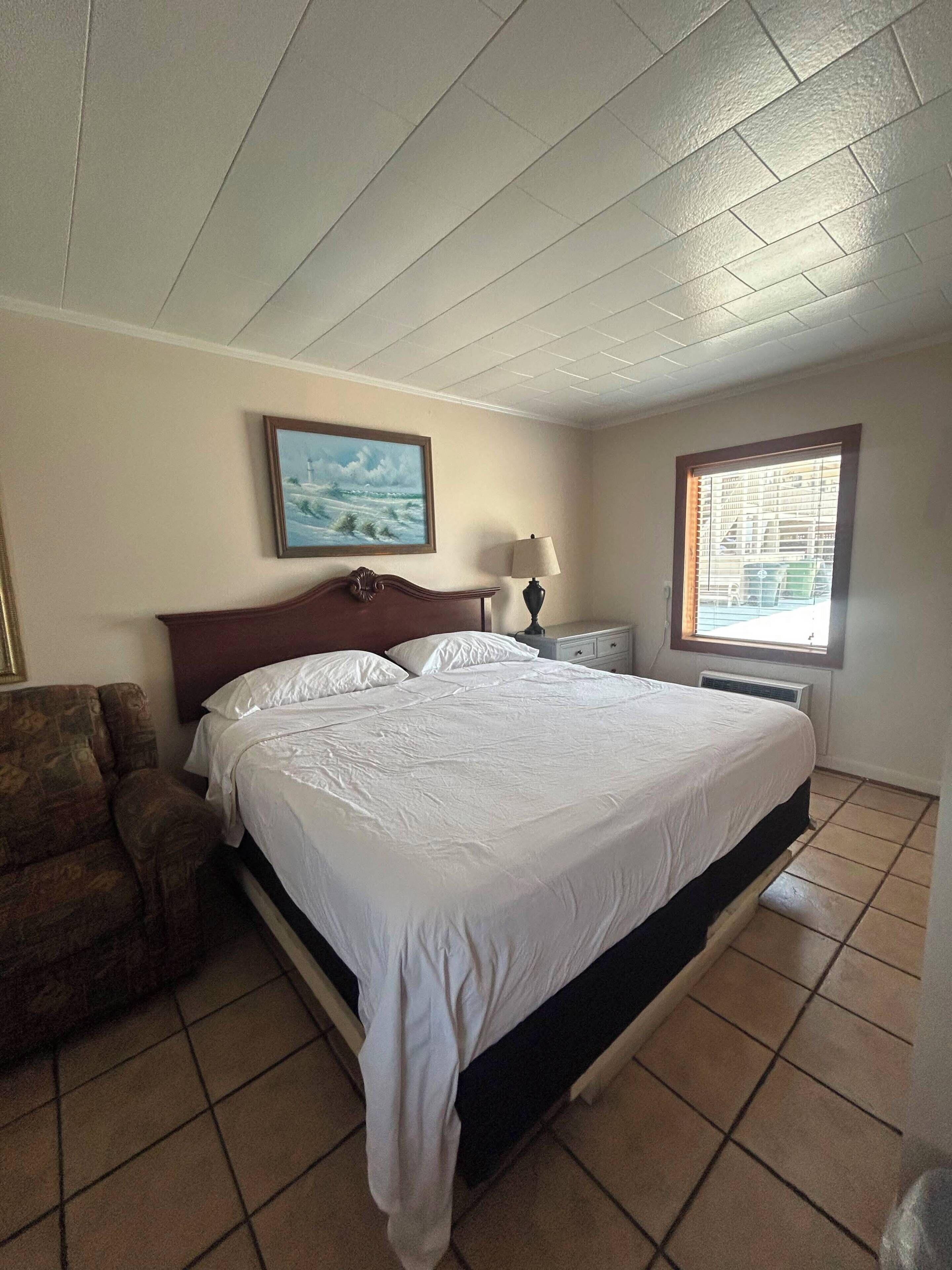 A hotel room with a bed, a chair, a nightstand, a lamp, a painting, and a window.