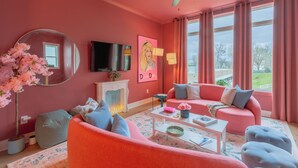A pink-themed living room with a ceiling fan, a flat-screen TV, a fireplace, and a large window with curtains. - Dolly's Pink Palace: Rooftop Deck (Nashville)