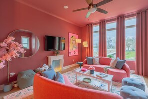 Ein in Rosa gehaltenes Wohnzimmer mit Deckenventilator, Flachbildfernseher, Kamin und einem großen Fenster mit Vorhängen.
