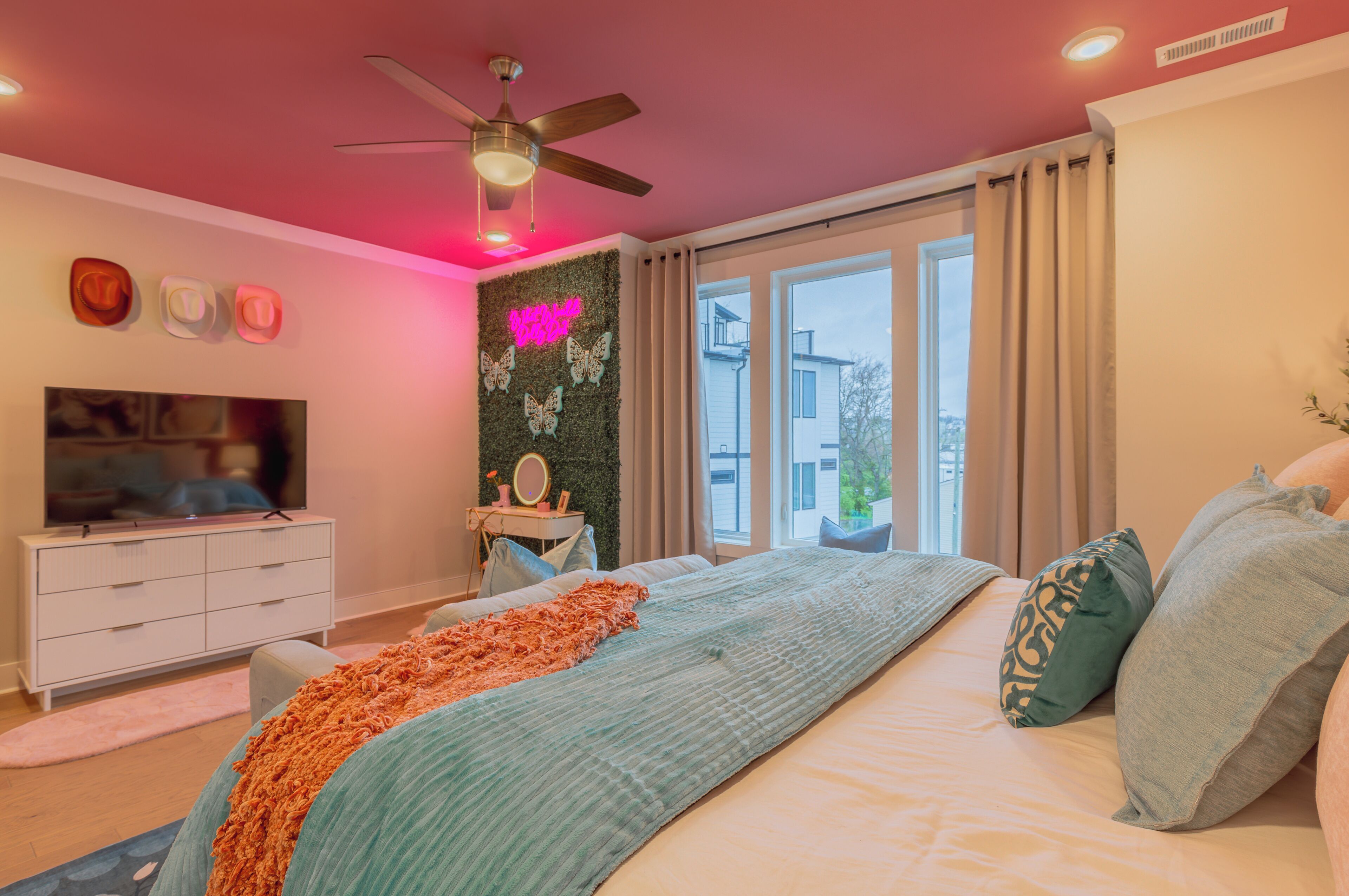 Een slaapkamer met een bed, plafondventilator, televisie, dressoir en raam met gordijnen.