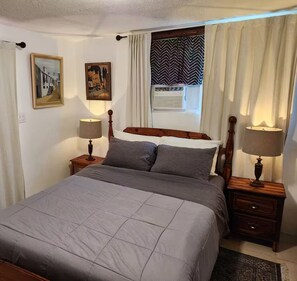 Ein ordentlich bezogenes Bett mit einer grauen Decke, zwei Nachttische mit Lampen, ein Fenster mit Vorhängen und zwei gerahmte Bilder an der Wand.