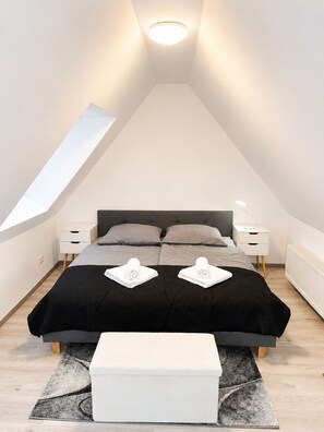 Ein modernes Schlafzimmer mit schrägen Wänden, einem Bett mit schwarzer Bettdecke, zwei Nachttischen mit Lampen und einer kleinen weißen Bank.