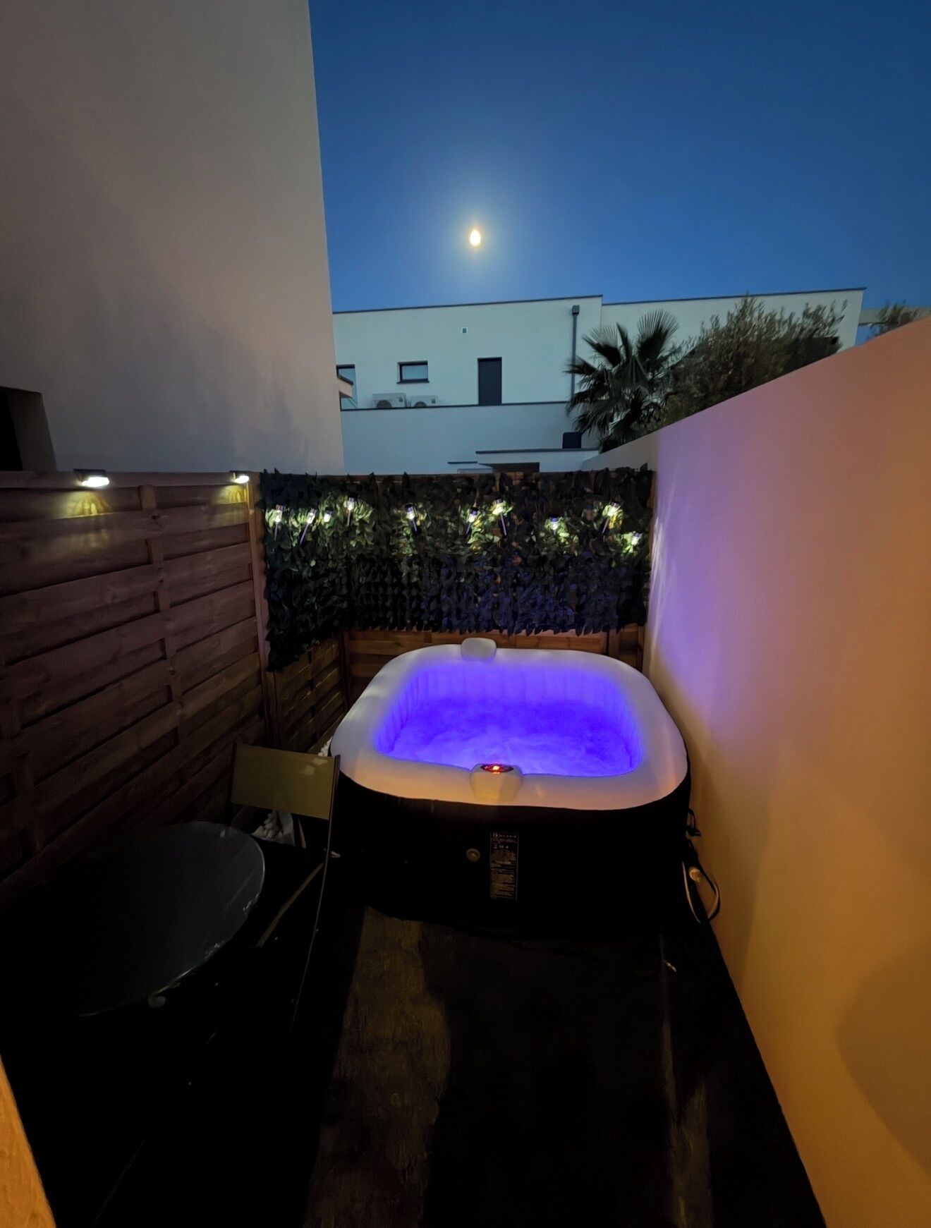 A hot tub with a blue light, surrounded by a wooden wall and a stone wall with plants.