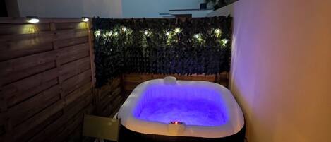 A hot tub with a blue light, surrounded by a wooden wall and a stone wall with plants.