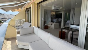 A balcony with a white sofa, a glass table, and a view of the city. - Spacious 3-Bedroom Apartment in a Naturist Village with Ocean Views (Agde)