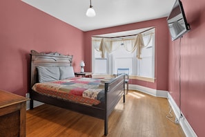A bedroom with a bed, a nightstand, a window with curtains, a television mounted on the wall, and a wooden floor. - Widder Haus, 1909 Historic Home with Porches & BBQ (Kansas City)