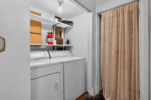 A laundry room with a washing machine, dryer, and ironing board. There are shelves above the appliances and a fire extinguisher on the wall. - Widder Haus, 1909 Historic Home with Porches & BBQ (Kansas City)