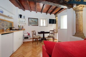 Eine kompakte Küche mit Holzbalken, ein Essbereich mit Stühlen, ein rotes Sofa und ein an der Wand befestigter Fernseher.