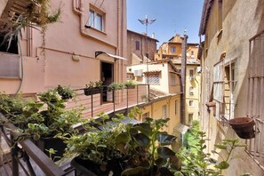 A narrow urban alley with buildings on either side, potted plants, and a balcony with railings.