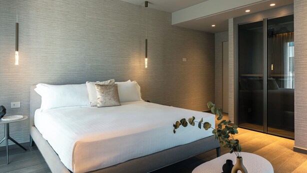 Ett modernt hotellrum med en stor säng, ett litet runt bord, en vas med en växt och en skjutdörrsgarderob.