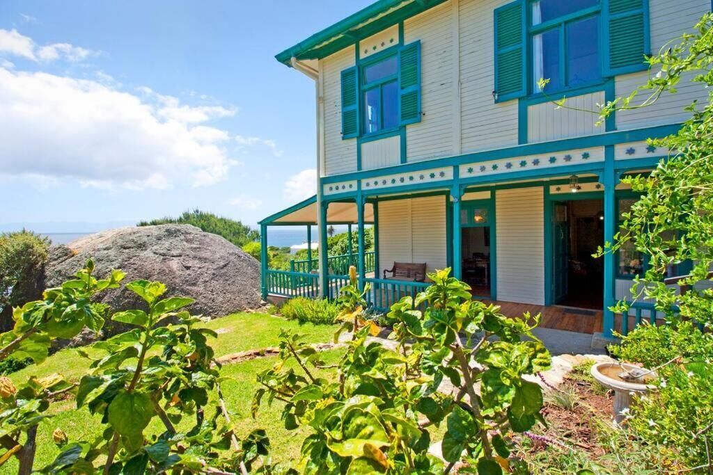 A two-story house with a porch, a large rock in the yard, and lush greenery.