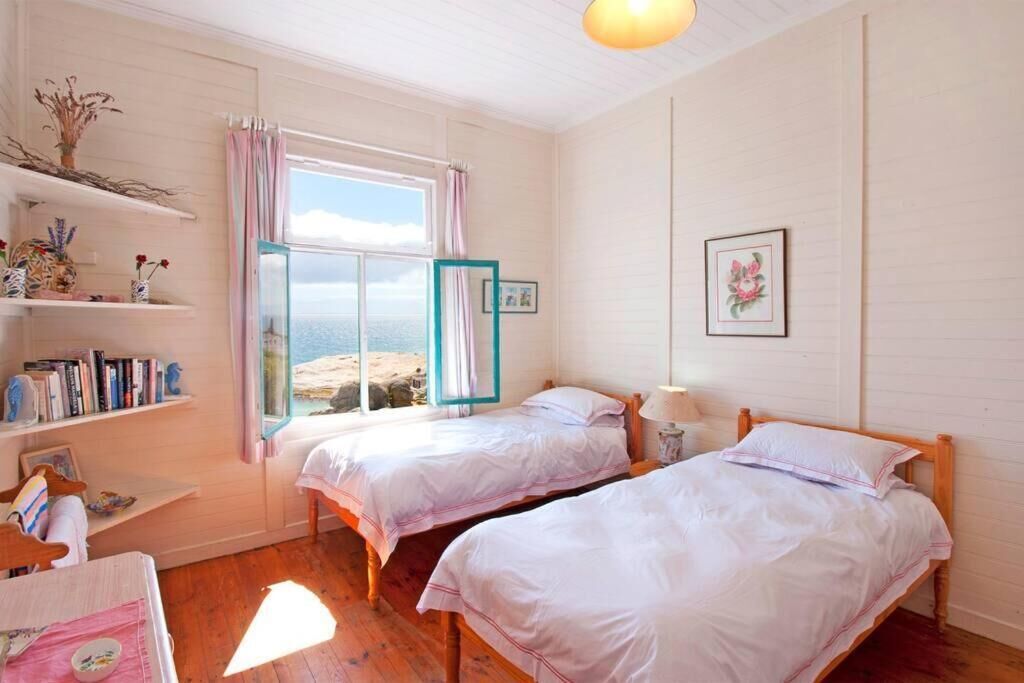 A bedroom with two beds, a window with curtains, a shelf with books, and a framed picture on the wall.
