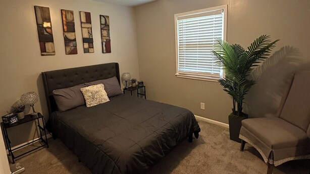 A bedroom with a bed, a nightstand, a chair, a window with blinds, a ceiling light, and wall art.