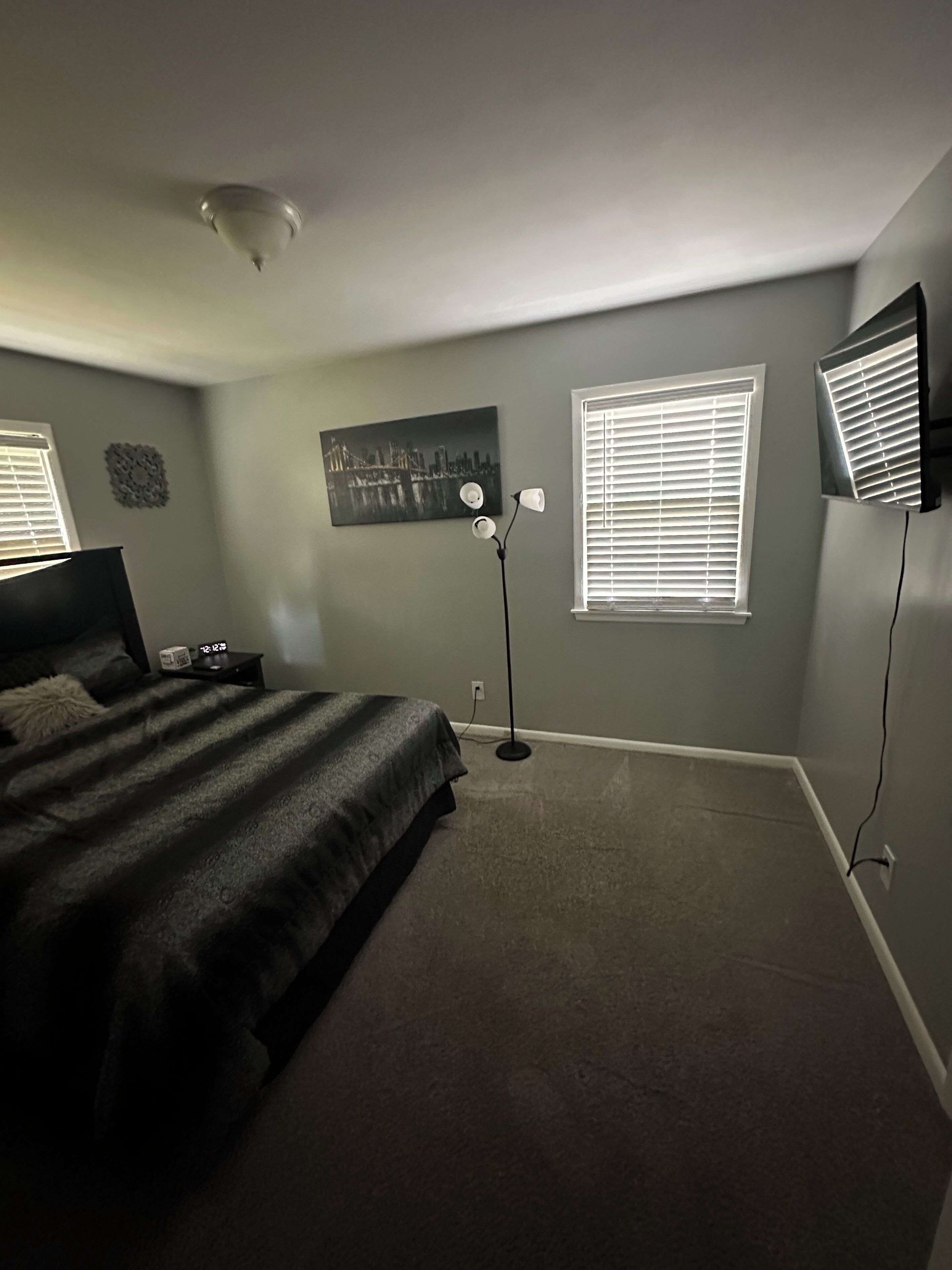 Une chambre à coucher comprenant un lit, une table de chevet, un téléviseur fixé au mur, une lampe sur pied et une fenêtre avec des stores.