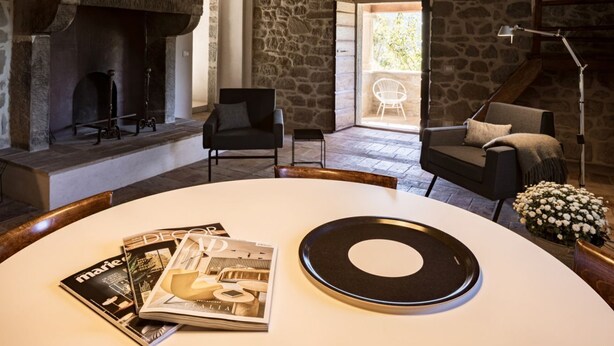 A room with a stone fireplace, a round table with magazines, a black armchair, and a white chair by a window. - Villa Bastia by MMega (San Leo Bastia)