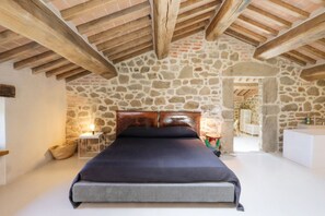 Ein Schlafzimmer mit Steinsandsteinwand, Holzbalkendecke, einem Bett, einem Nachttisch und einem Beistelltisch.