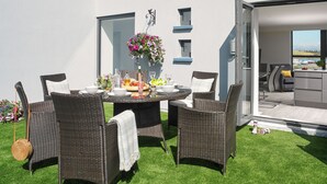 Un área de comedor al aire libre con mobiliario de mimbre, una mesa dispuesta con platos, vasos y una cesta de flores colgante.