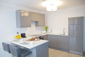 A modern kitchen with grey cabinets, a built-in oven, a refrigerator, and a dining area with a chair and table. - Asgard Apartments (Westport)