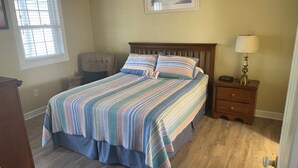 Quarto com cama, uma cadeira, mesa de cabeceira, abajur e janela.
