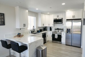 A modern kitchen with white cabinets, a marble island, black bar stools, stainless steel appliances, and a window with blinds. - Spacious Central San Diego Home! (San Diego)