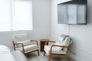 Ein modernes Zimmer mit einem Bett, zwei Sesseln, einem kleinen runden Tisch und einem an der Wand befestigten Fernseher.