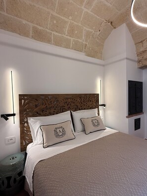 A bedroom with a wooden headboard, two pillows with monogrammed covers, a bedside lamp, and a wall-mounted TV. - Angel Apartments by Monholiday (Monopoli)
