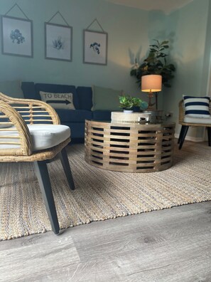 Ein Wohnzimmer mit einem Korbsessel, einem runden Holztisch, einem blauen Sofa und einer Wand mit gerahmten Kunstwerken.