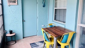 Ein Zimmer mit blauer Tür, einem Holztisch, gelben Stühlen, einem kleinen runden Tisch mit Pflanze und einem Fenster mit Rollläden.