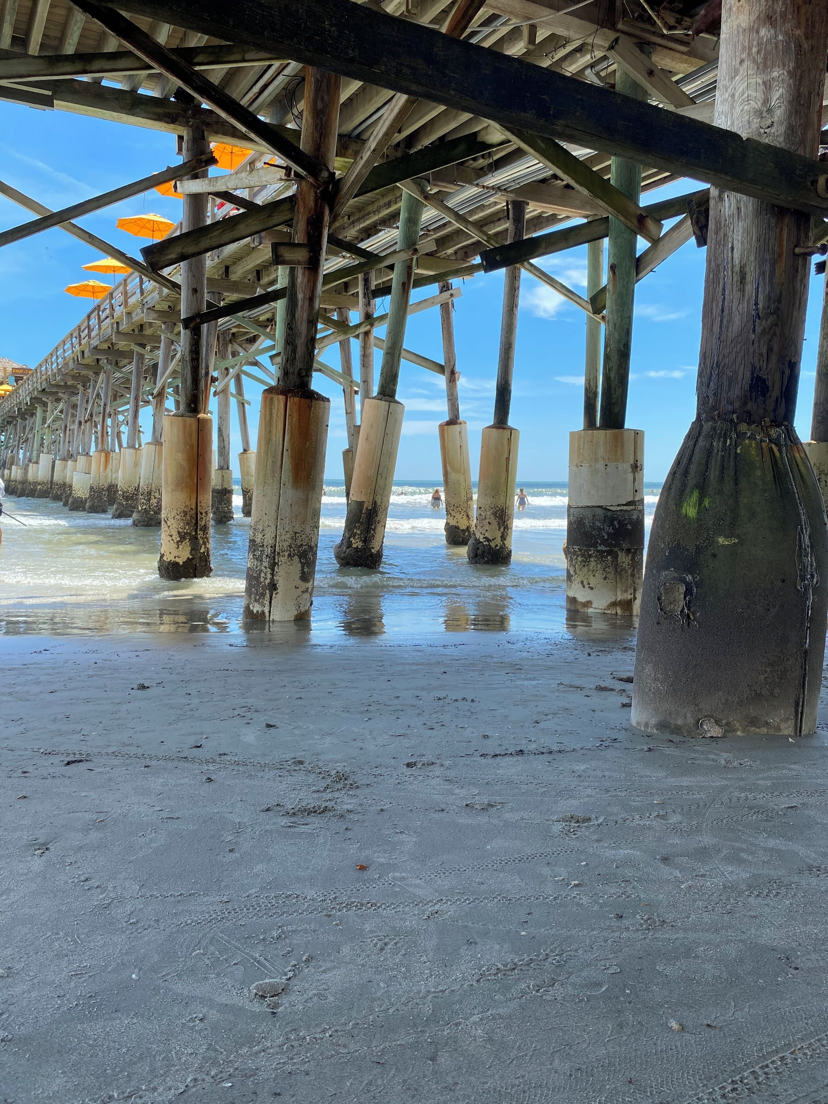 Una estructura de muelle con soportes de madera que se adentran en el agua, una playa arenosa y un cielo despejado.