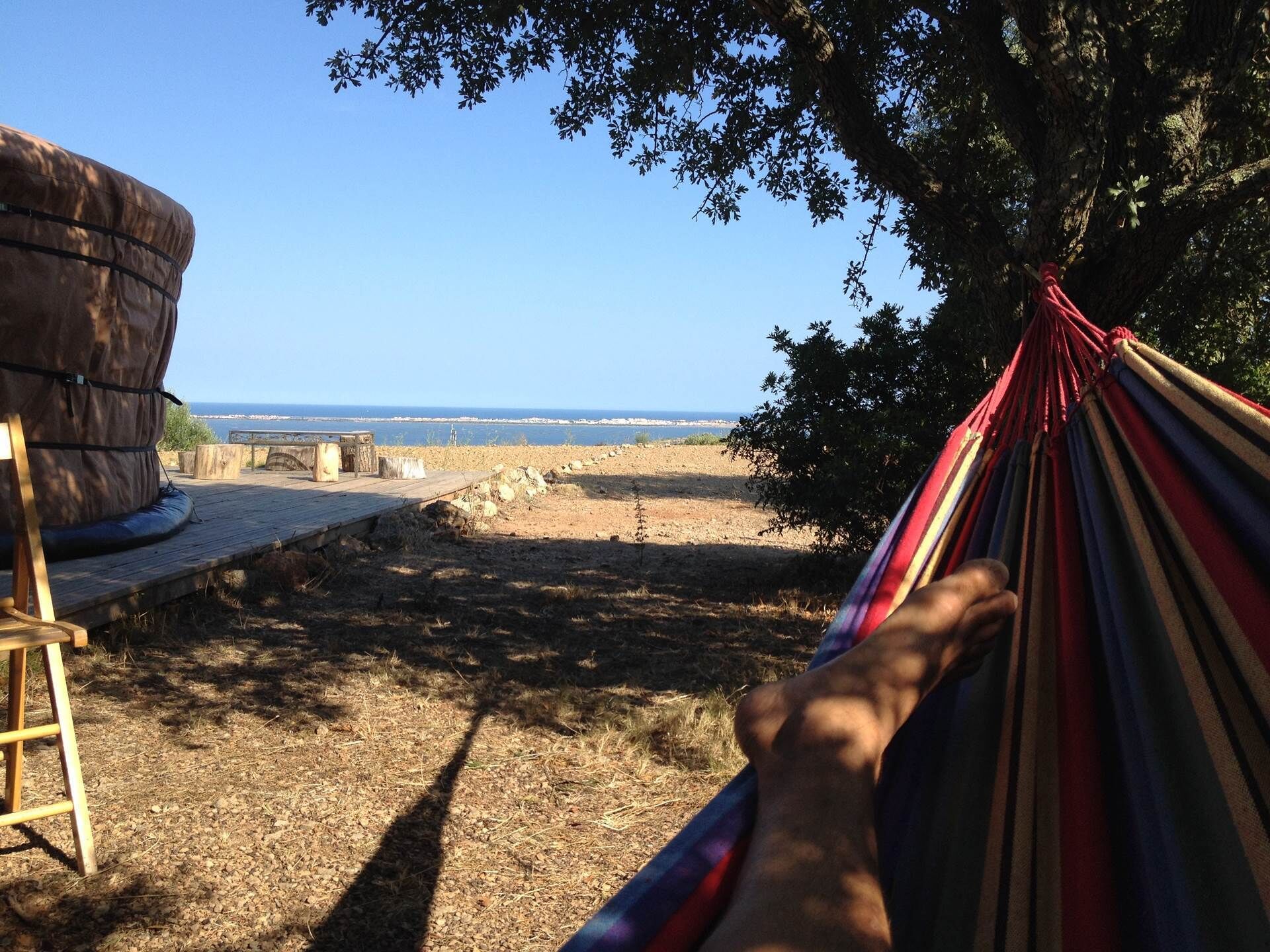 Un hamac est suspendu entre deux arbres, offrant une vue sur une plage, avec une terrasse en bois et une grande formation rocheuse.