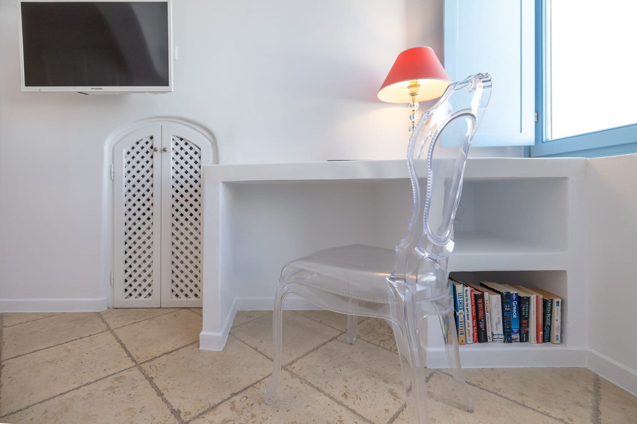 A room with a clear plastic chair, a white shelf with books, a red lamp, and a TV on the wall.