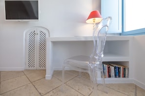 A room with a clear plastic chair, a white shelf with books, a red lamp, and a TV on the wall. - Ardelion Suite, Divine Sea Views, Outdoor Jacuzzi, Santorini (Santorini)