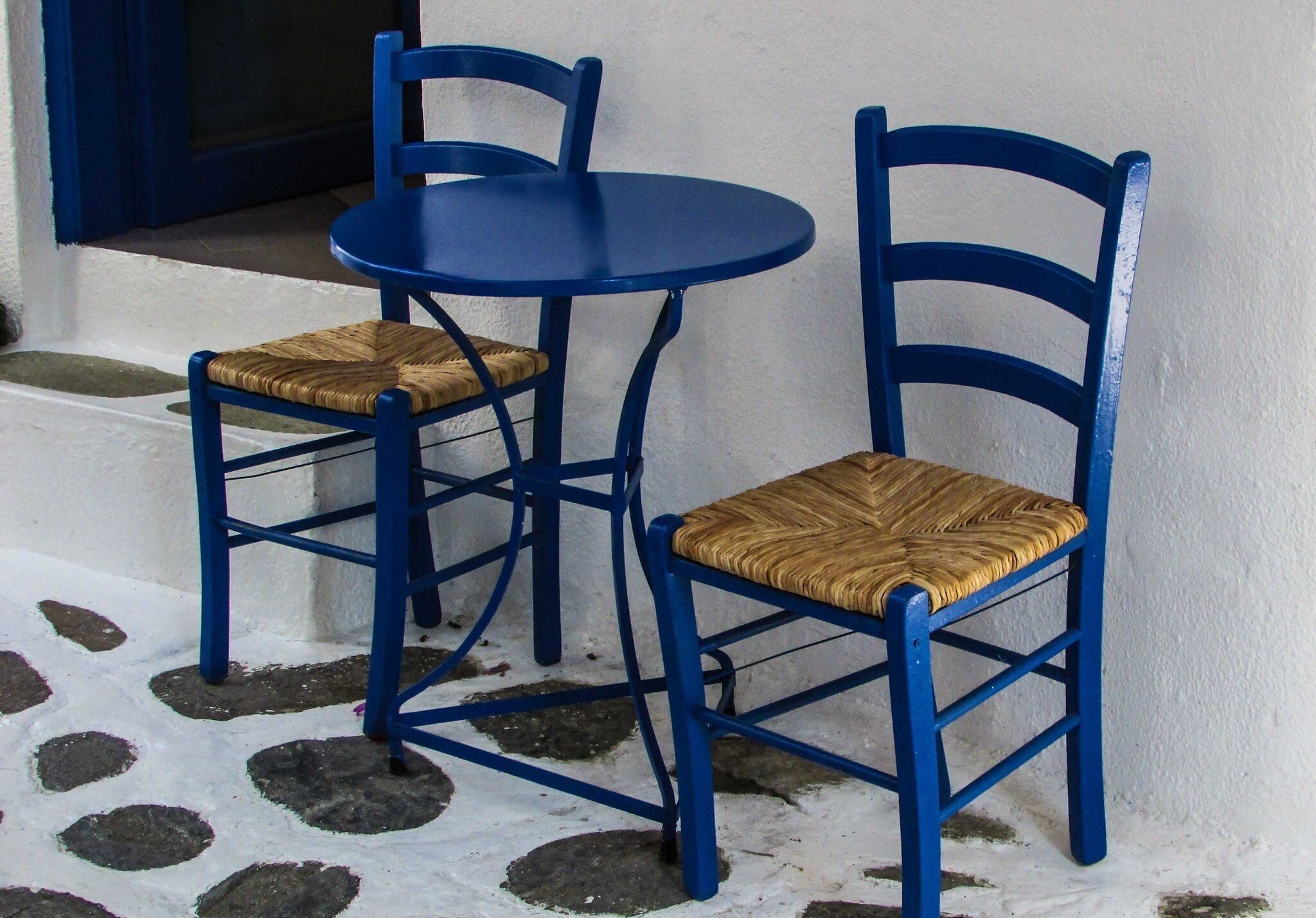 Twee blauwe stoelen met gevlochten zitting en een kleine ronde tafel daartussen, geplaatst tegen een witte muur met een vloer van steenmotief.