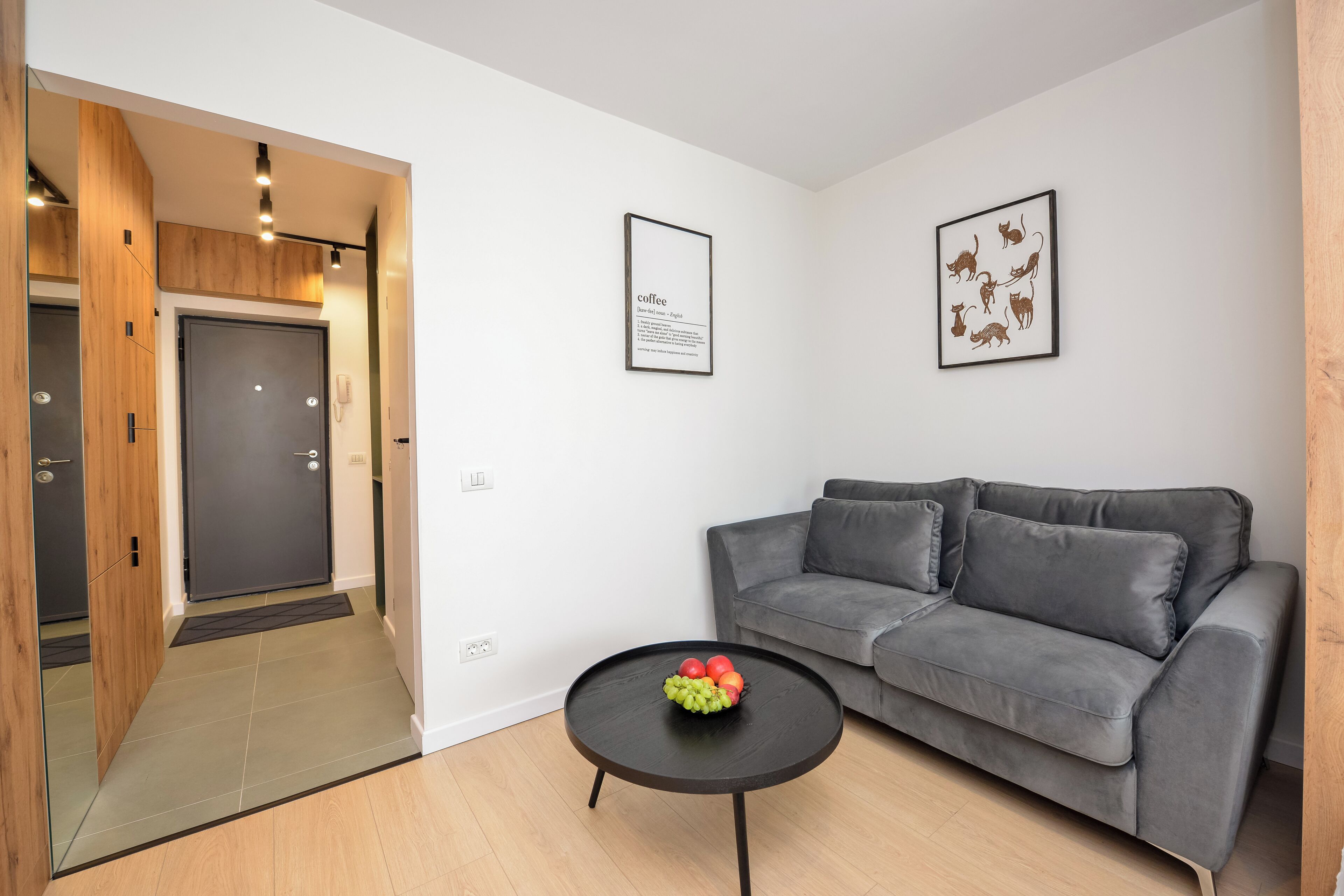 Una sala de estar moderna con un sofá gris, una mesa de centro negra con fruta y un cuadro de gatos en la pared.