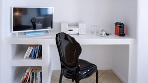 Ein weißer Schreibtisch mit einem Computer, Lautsprechern und einer Kaffeemaschine. Davor steht ein schwarzer Stuhl. Unter dem Schreibtisch befindet sich ein Bücherregal.