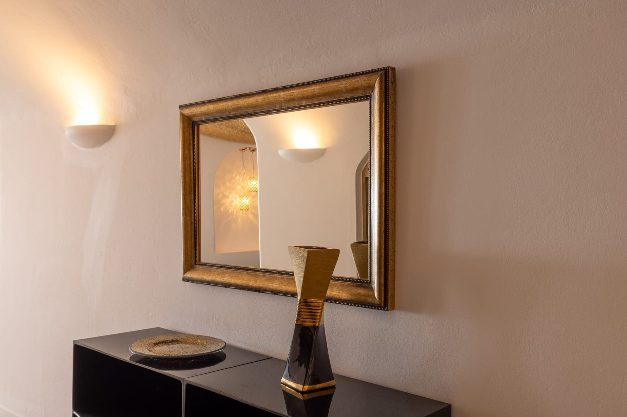 Een spiegel aan de muur met gouden lijst, een vaas en twee wandlampen.