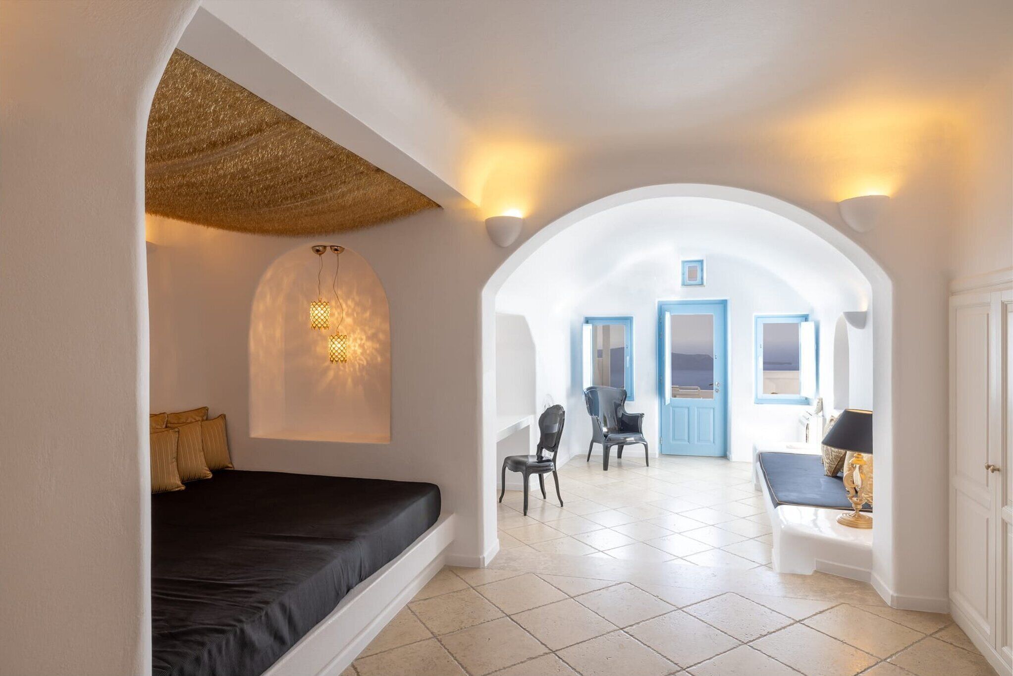 Een hotelkamer met een bed, een zithoek met een stoel en een tafel, en uitzicht door een boogvormige deuropening.