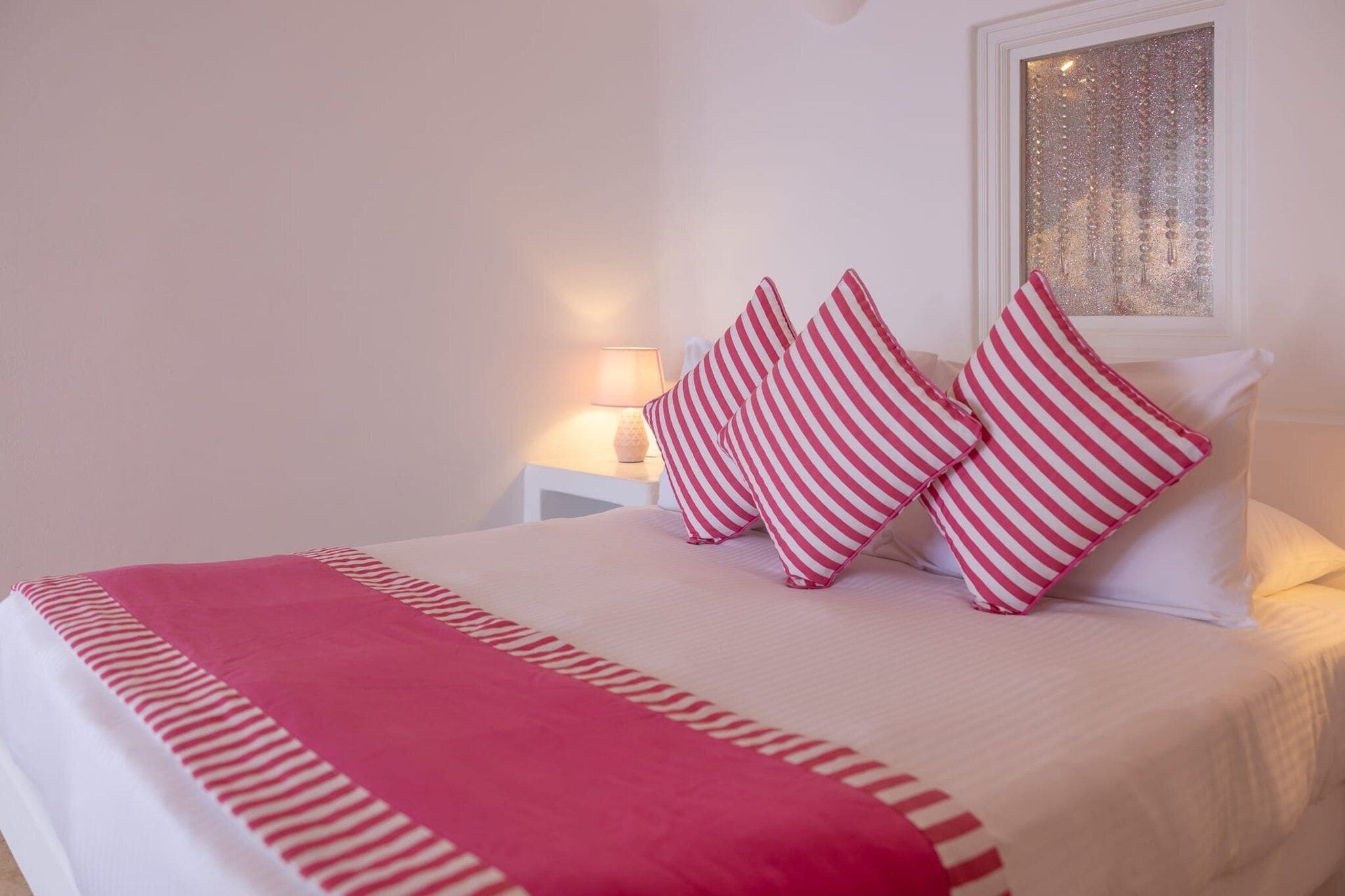 Un lit bien fait, avec des oreillers à rayures et une couverture rose, une lampe de chevet et une fenêtre agrémentée d’un rideau décoratif.
