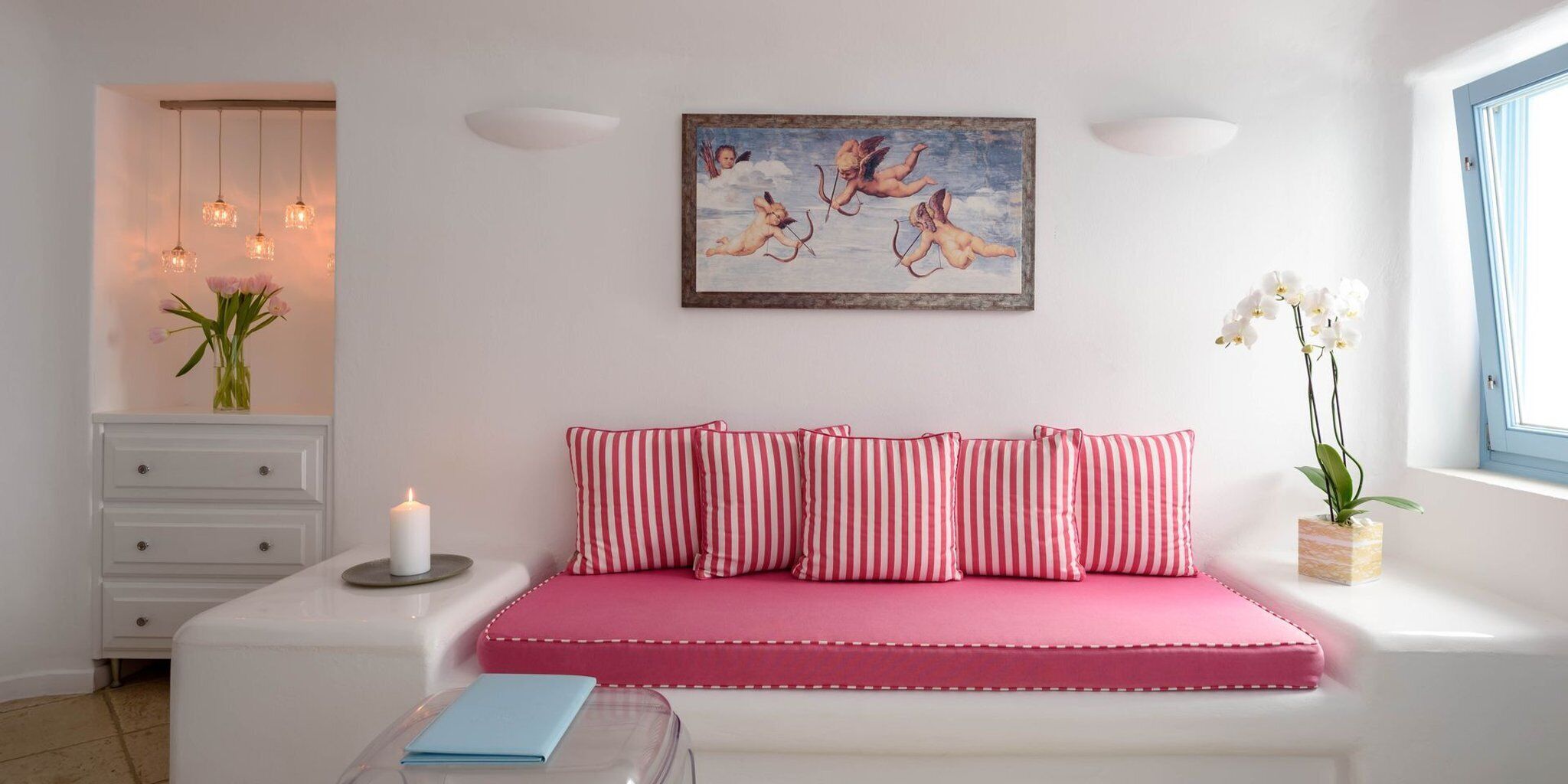 Une chambre avec un lit rose, des coussins à rayures, un tableau représentant des anges, un vase de fleurs et une fenêtre avec des rideaux.