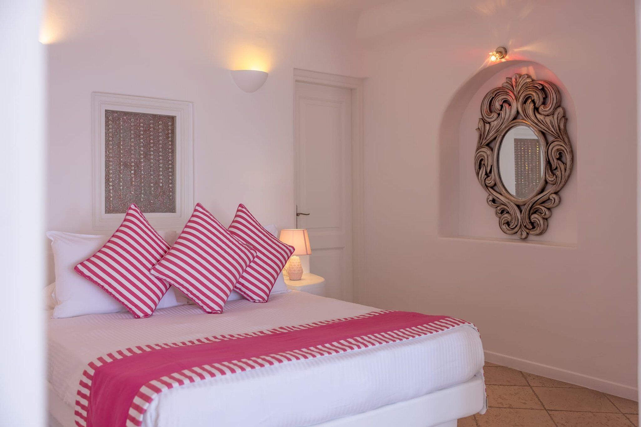 Een slaapkamer met een bed, kussens, een nachtkastje, een lamp, een spiegel en een raam met decoratieve lijst.