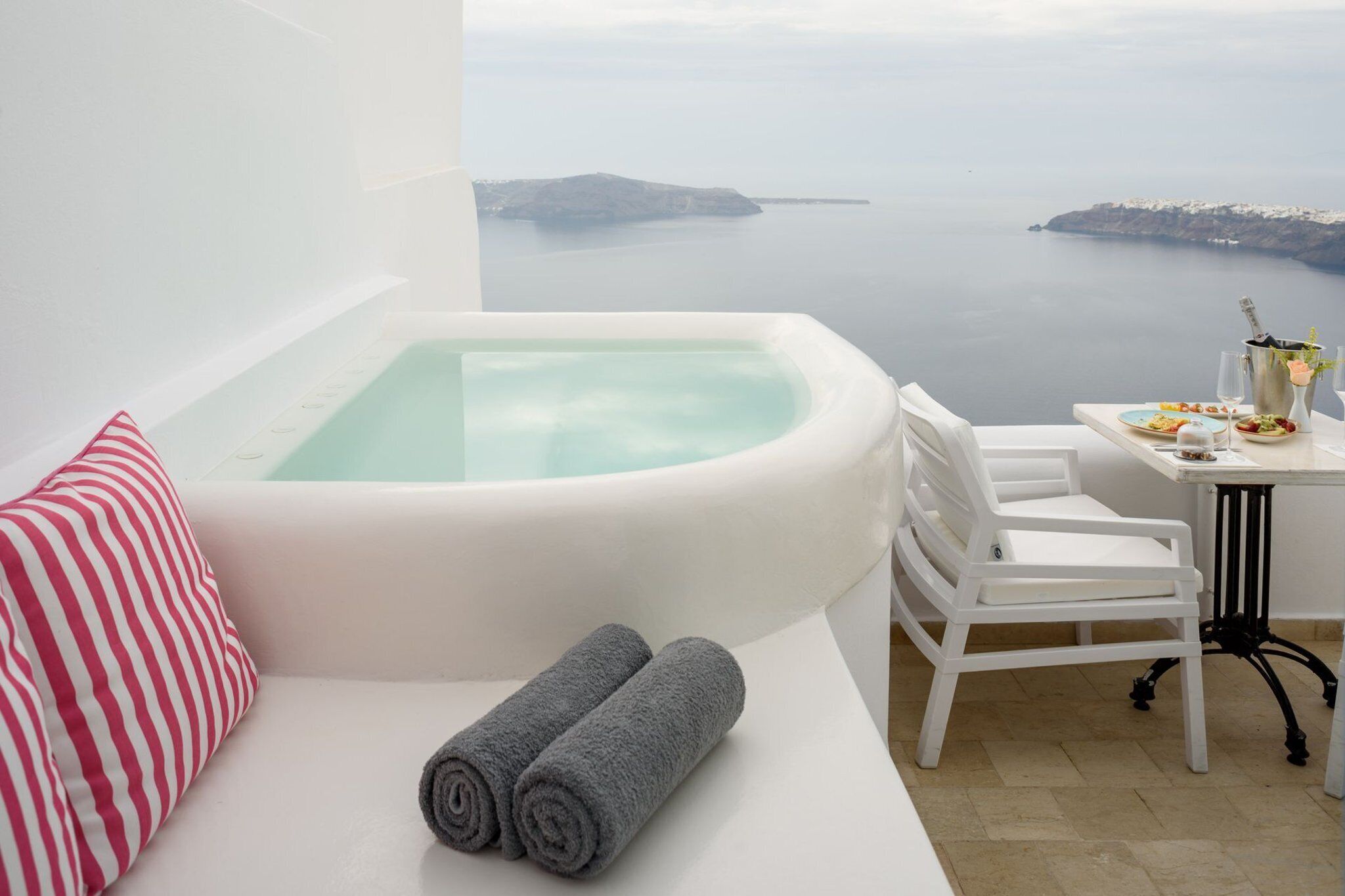 Un balcon avec un jacuzzi, un canapé blanc, un coussin à rayures, deux serviettes roulées et une table sur laquelle se trouvent une assiette de nourriture et une boisson.