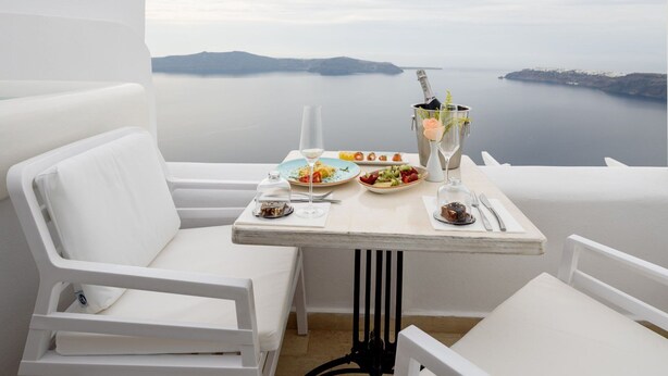 Un balcó blanc amb taula preparada per a dinar, amb vista a un mar tranquil i a la terra lluny.