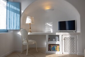 A room with a white desk, a television, a white chair, a bookshelf with books, and a window with blue shutters. - Coralyon Suite, Divine Sea Views, Outdoor Jacuzzi, Santorini (Santorini)