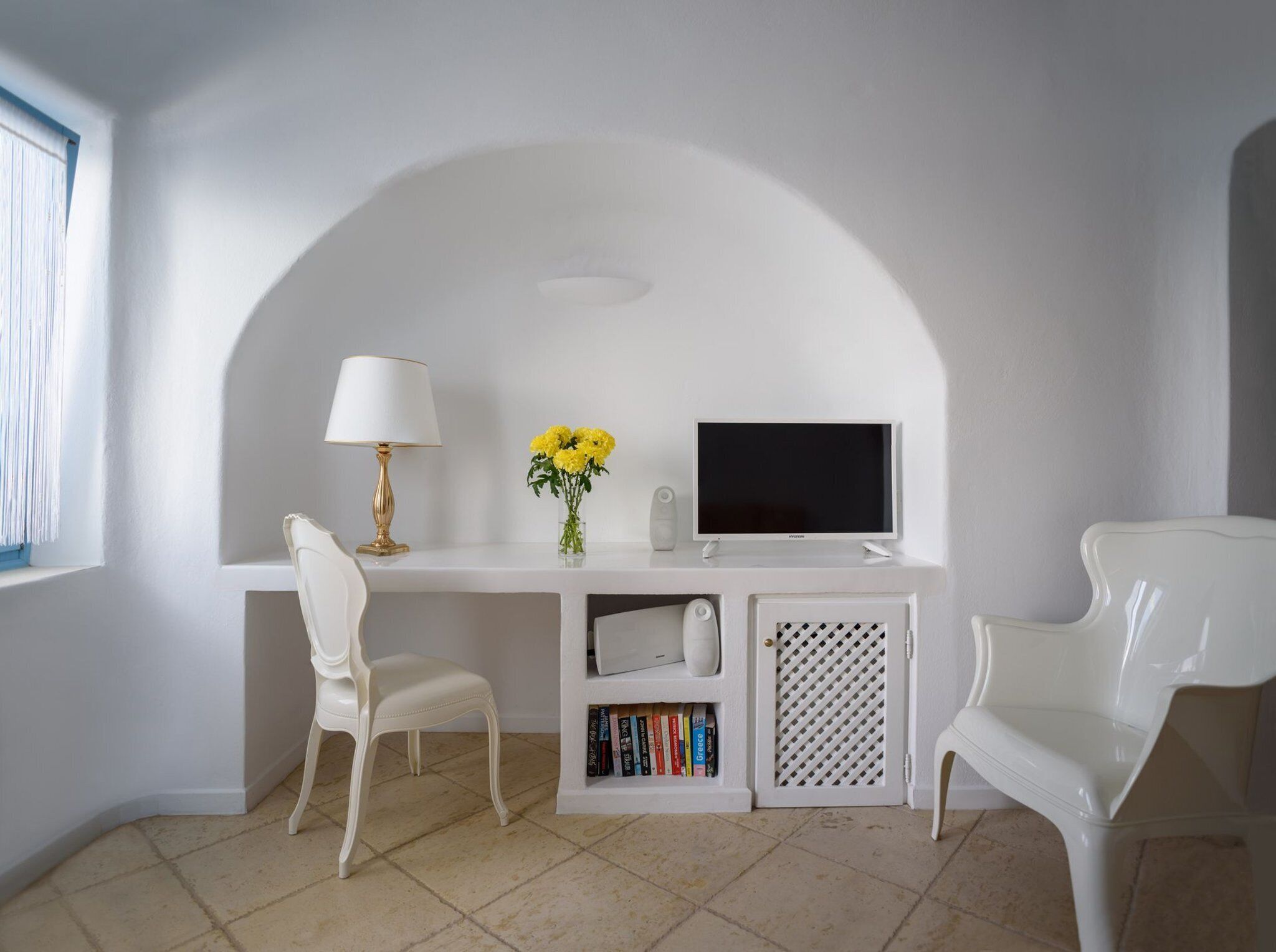 A white study area with a desk, two white chairs, a lamp, a vase with flowers, a TV, and a bookshelf.